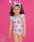 Young girl smiling wearing the RuffleButts + RuggedButts - Girls Coastal Breeze Floral Bow V-Back One Piece Swimsuit: Pink with floral print and ruffled striped sleeves, styled with a matching striped bow headband against a bright pink background in a curated children’s boutique setting.