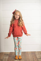 A young girl with blonde hair wearing a rust-orange ruffle-hem hoodie, light blue floral leggings, and brown sandals, standing against a white shiplap wall.