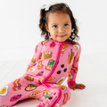Toddler girl wearing a colorful Kiki + Lulu - Pink Breakfast Convertible Footie featuring playful breakfast food prints, sitting on a white floor in soft natural light.