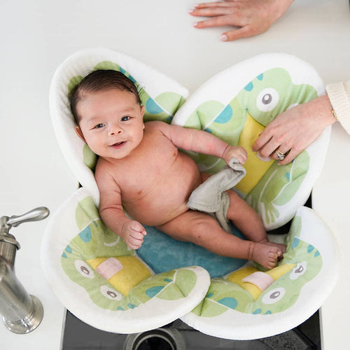 Baby lying comfortably in a soft frog-shaped bath cushion while being gently supported by adult hands, showcasing Blooming Baby - Blooming Bath Pond Pal: Frog from a curated children’s boutique.