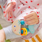 Baby wearing pink floral pajamas holding a colorful Rattlefish Wooden Baby Rattle, a curated children’s boutique item perfect for sensory play and mom life essentials.