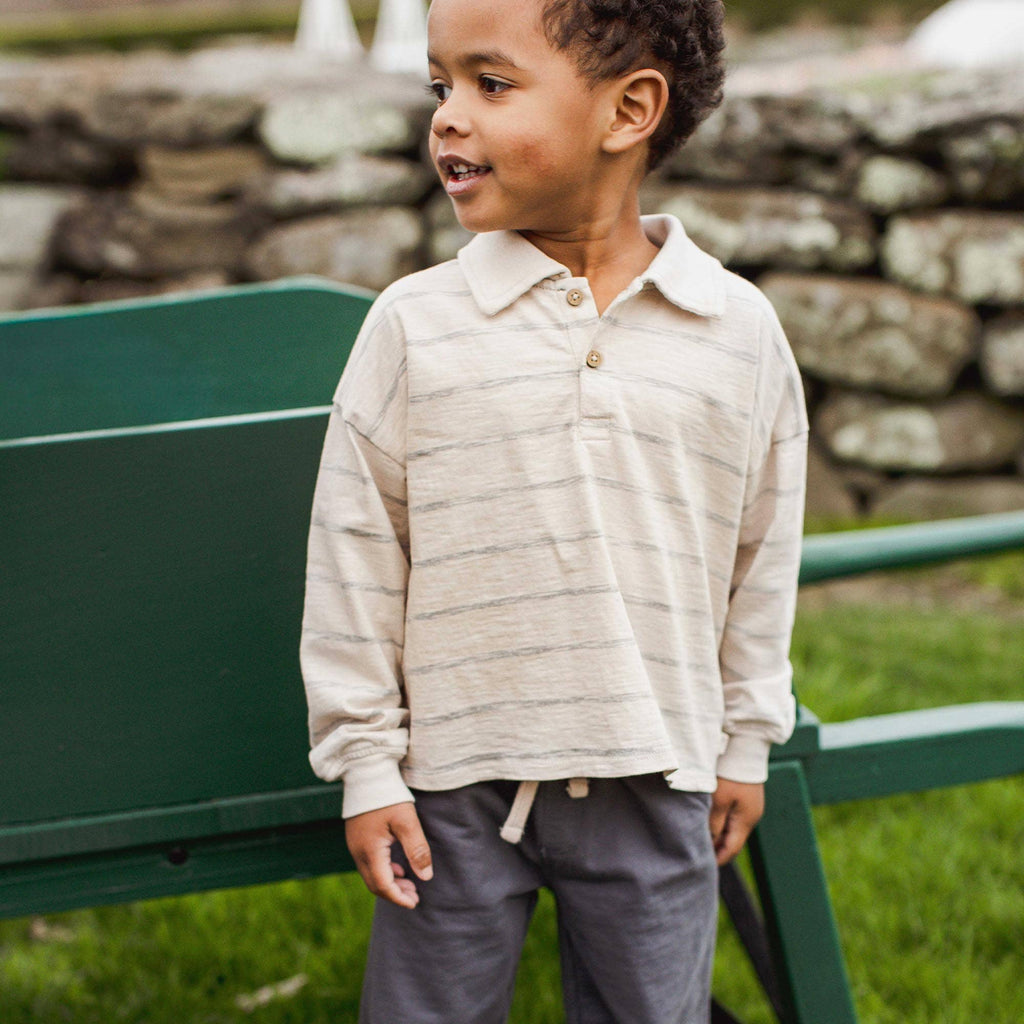 A young child wearing a cream and gray striped long-sleeve polo shirt and dark gray pants, standing outdoors next to a green bench with a stone wall in the background.