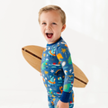 Young boy smiling and holding a wooden surfboard while wearing Kiki + Lulu - Blue Dino Kid Pajamas with colorful dinosaur and tropical prints, styled for cozy and playful comfort.