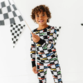 Child wearing Kiki + Lulu - Racecar Kids Pajamas with colorful racecar and checkered flag pattern, holding a checkered flag in a bright, playful setting.