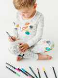 Child sitting and coloring designs on Bee Colorful Co. - Space Coloring Pajamas 2 piece set made from Bamboo, featuring space-themed patterns with markers scattered around.