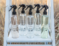 Five clear spray bottles labeled Lemon & Lavender, La Belle, White Tea, Oatmeal Milk & Honey, and Sunday Beach arranged on a white wooden shelf outdoors, each featuring Simplified Soap - Body Spray.