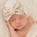 Sleeping newborn baby wearing the ilybean - GOLDIE Bow Newborn Girl Hospital Hat with a pearl-embellished satin bow in soft neutral tones, cozy and stylish baby accessory.