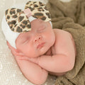 Newborn peacefully sleeping wearing a white headband adorned with an ilybean - LUXE LEOPARD BOW, showcasing stylish kids clothing in a cozy, neutral-toned setting.