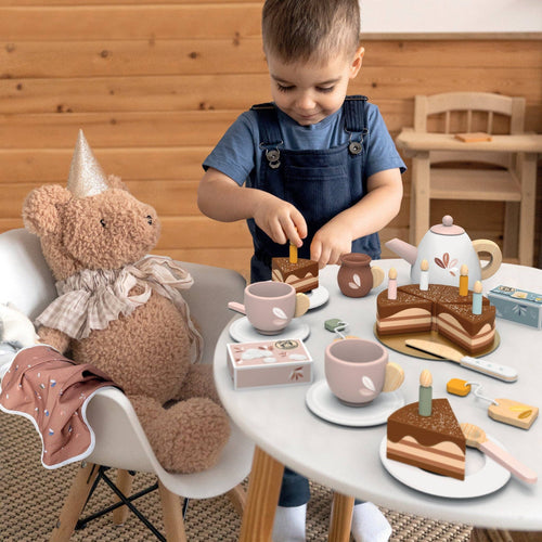 Child playing with the Speedy Monkey - Birthday Tea 18 Piece Pretend Play Set, serving wooden cake slices and tea on a small white table with a teddy bear guest in a cozy, stylish children’s boutique setting.