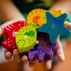 Close-up of colorful wooden ocean-themed magnetic game pieces held in a child's hands, featuring a starfish, fish, and crab from My Very First Games - Here, Fishy, Fishy! Magnetic Game.