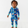 Toddler wearing the Kiki + Lulu - Cozy & Courageous Superhero Convertible Footie in a bright blue fabric with colorful superhero and star patterns, featuring teal cuffs and zipper trim.
