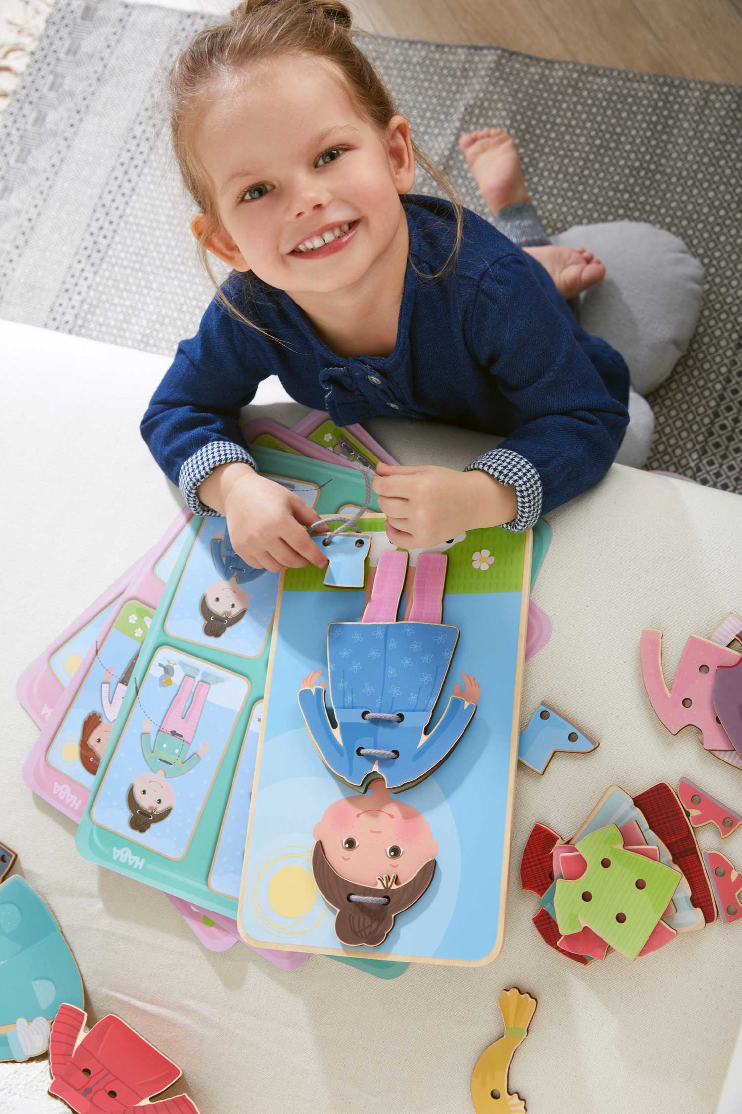 A young girl is smiling while playing the Dress Me Threading & Lacing Game, practicing fine motor skills with colorful wooden pieces on a soft, neutral-toned rug.
