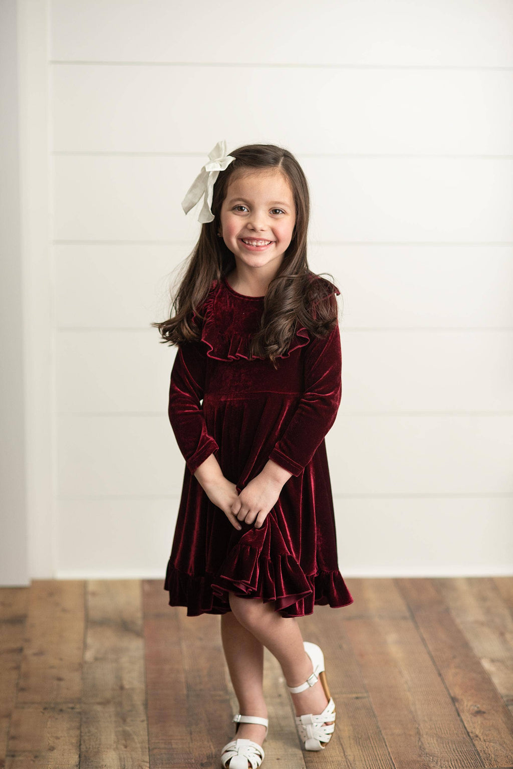 Young girl wearing a burgundy velvet dress with a white bow in her hair, standing on a wooden floor.