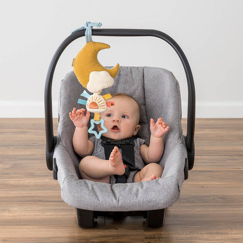 Baby sitting in a gray infant car seat reaching for the Itzy Ritzy - Bitzy Notes™ Musical Pull-Down Toy Cloud/Sun hanging from the handle, showcasing a soft neutral and playful design.