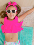 Young girl lying on a turquoise towel wearing RuffleButts + RuggedButts - Girls Tropical Resort Flounce Bikini 2-Piece Swimsuit with Signature Rear Ruffles: White, white heart-shaped sunglasses, and a bright pink headband with a bow, smiling cheerfully in a cozy and playful setting.