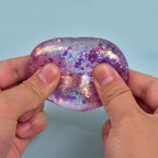 A person's hands holding a translucent, heart-shaped gel toy filled with purple and pink glitter against a light blue background.