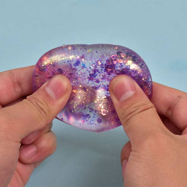 A person's hands holding a translucent, heart-shaped gel toy filled with purple and pink glitter against a light blue background.