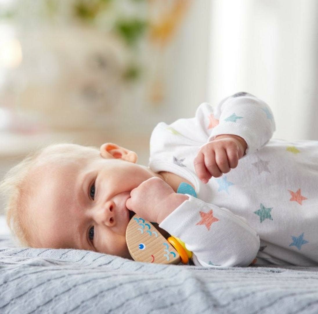 Baby lying on a soft gray blanket chewing on a Rattlefish Wooden Baby Rattle, wearing a white outfit with colorful star patterns, showcasing cozy baby essentials for modern families.