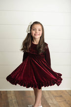 Young girl wearing a burgundy velvet dress with a white bow in her hair against a white background.