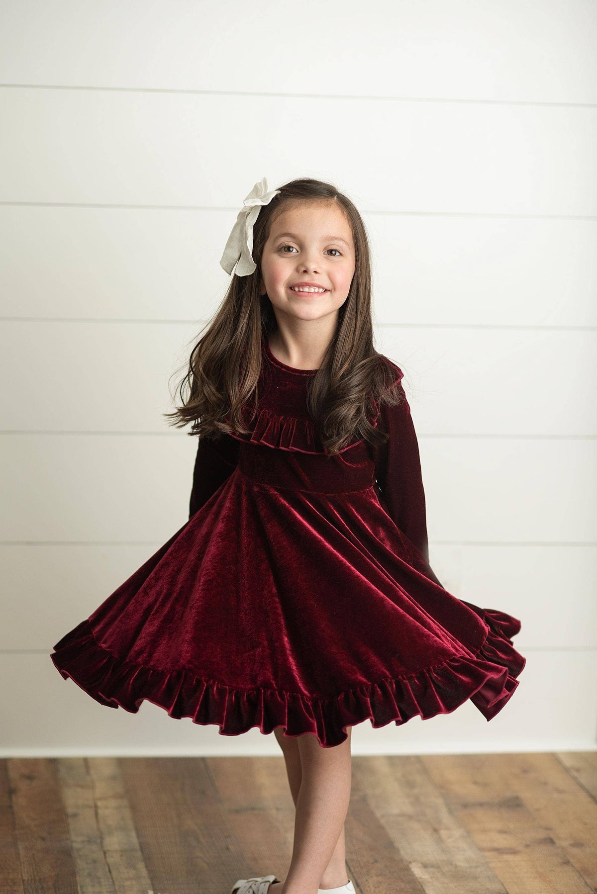 Young girl wearing a burgundy velvet dress with a white bow in her hair against a white background.