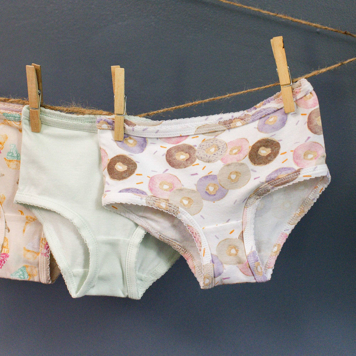 Three pairs of girl brief underwear hanging from wooden clothespins on a rope against a dark gray wall. The front pair is white with a pastel donut print, and the pair behind it is a solid pale mint green.