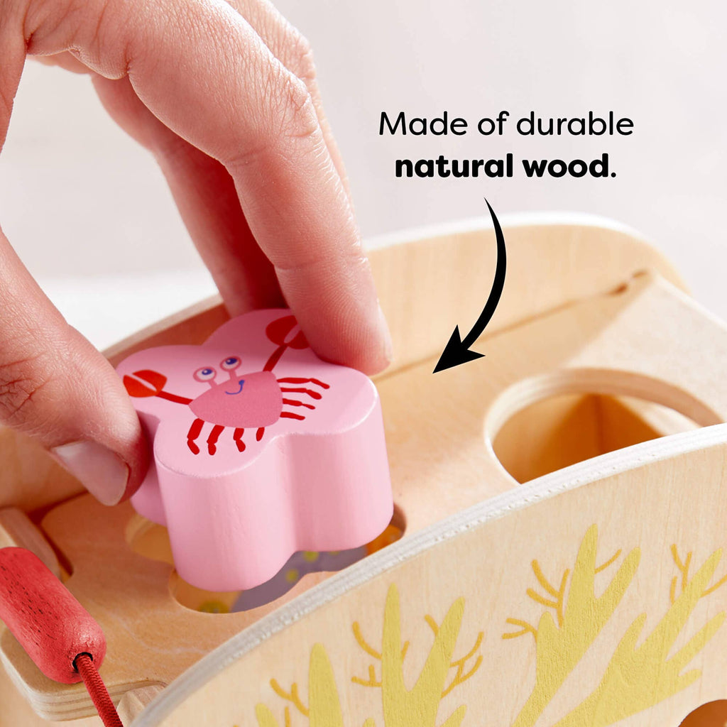 Hand placing a pink crab-shaped wooden piece into the matching hole of the Underwater World Wooden Shape Sorter made of durable natural wood, promoting educational play.