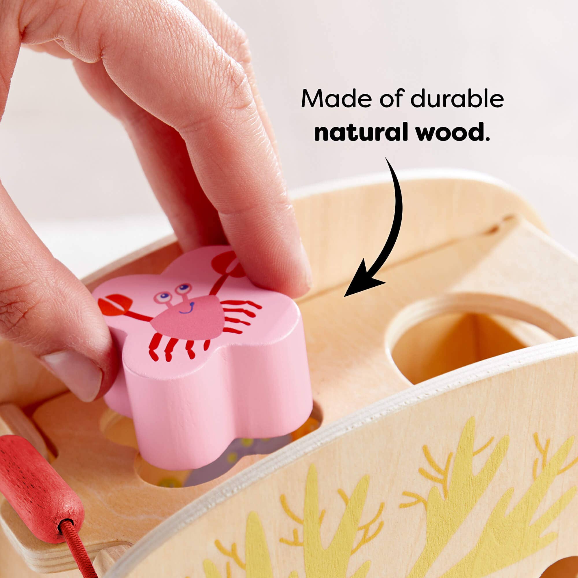 Hand placing a pink crab-shaped wooden piece into the matching hole of the Underwater World Wooden Shape Sorter made of durable natural wood, promoting educational play.