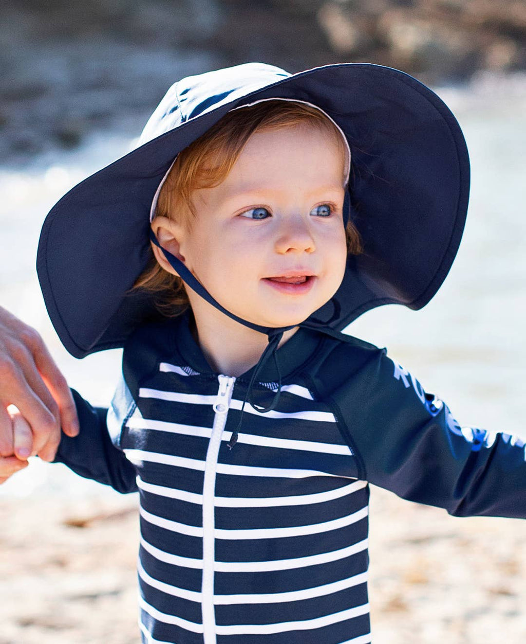 Toddler wearing a navy and white striped swim shirt and a navy wide-brimmed RuffleButts + RuggedButts - Kids Sun Protective Hat, holding an adult's hand by a sunny beach.