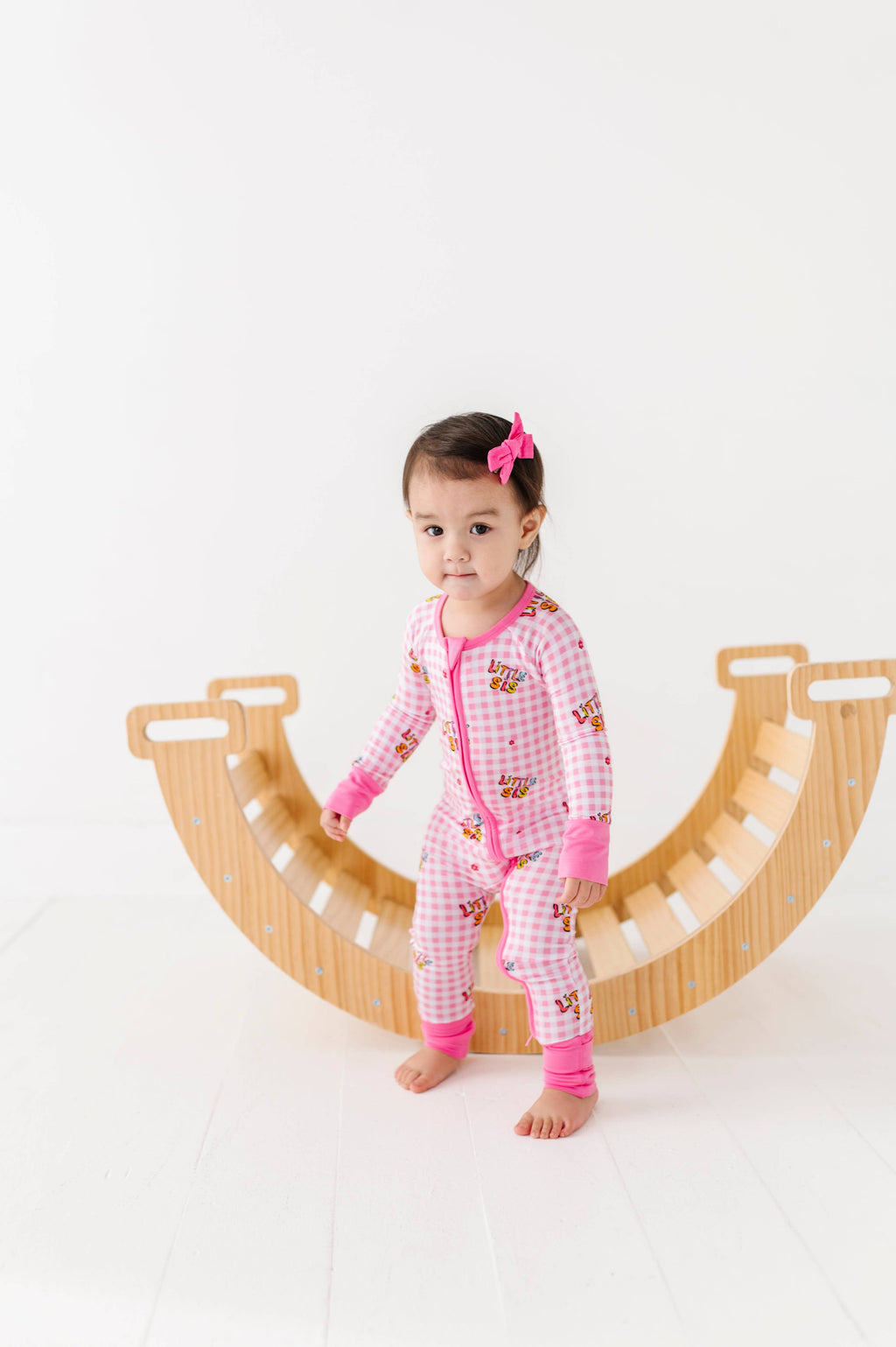 Child wearing a pink checkered onesie with cartoon characters, standing on a wooden arch.