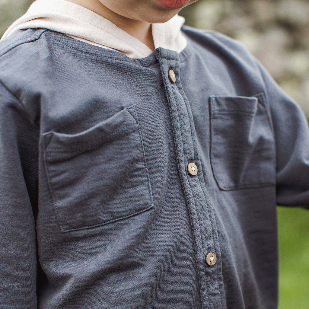 Close-up of a child wearing a dark slate blue button-up shirt with a white hood, featuring two chest pockets and wooden buttons.