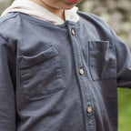 Close-up of a child wearing a dark slate blue button-up shirt with a white hood, featuring two chest pockets and wooden buttons.