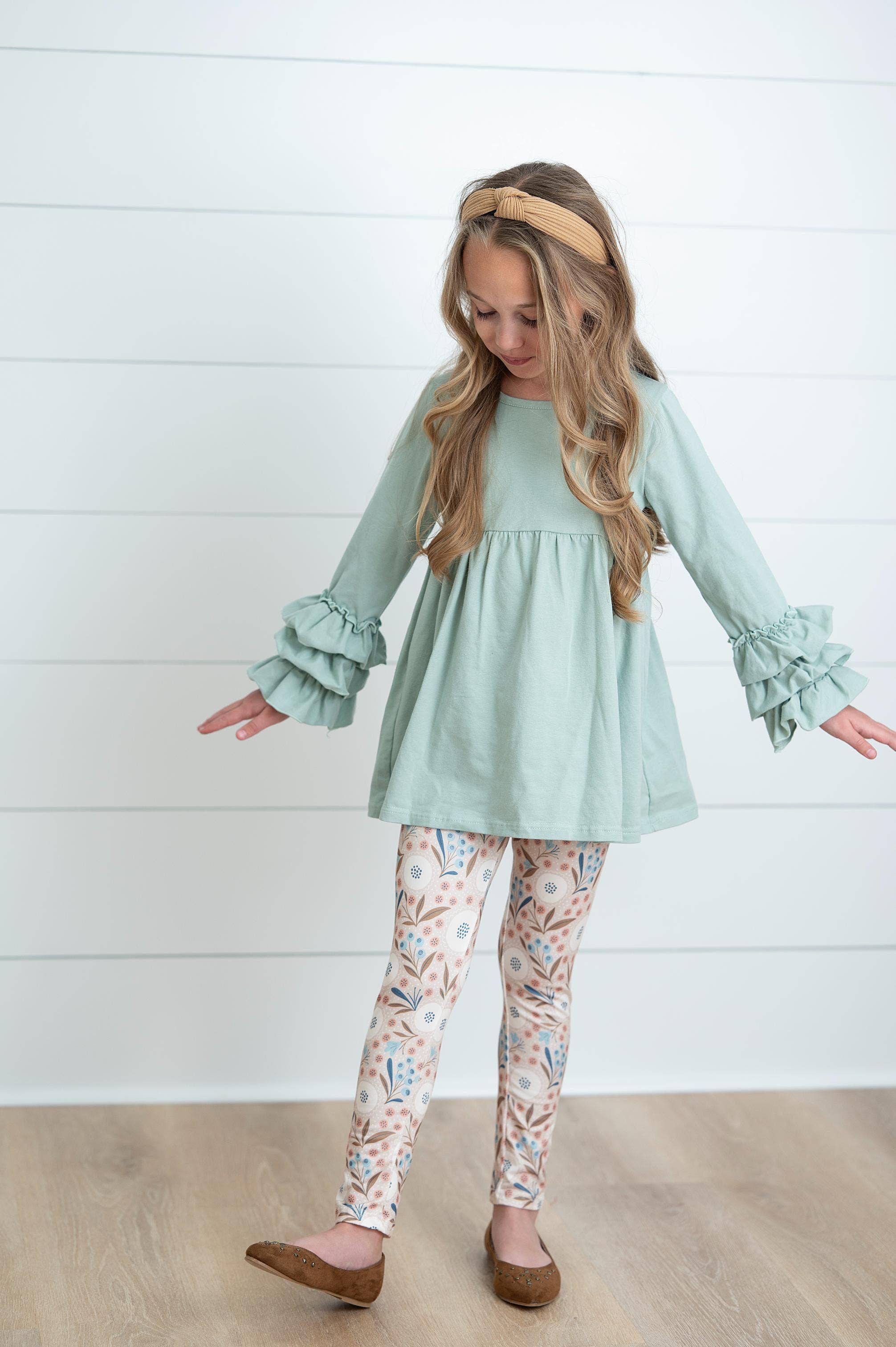Young girl wearing a light green top and patterned pants standing against a white wooden panel background.