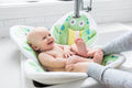 A happy baby relaxing in a soft, green frog-themed bath seat inside a white sink, showcasing the Blooming Baby - Blooming Bath Pond Pal: Frog from a curated children’s boutique.