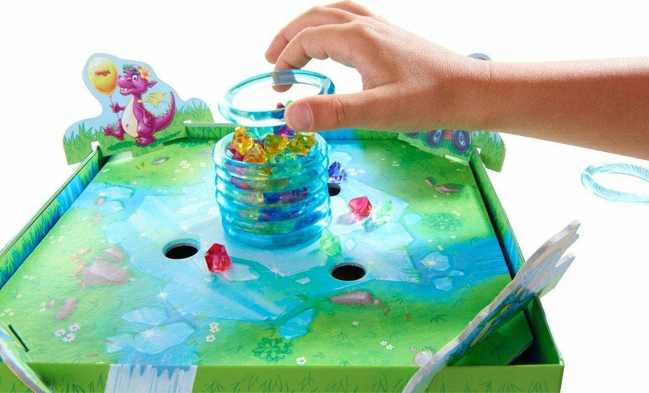 Hand reaching to play with colorful translucent dragon gems in the center of the Dragon's Breath Game, set on a vibrant green game board with whimsical illustrations.