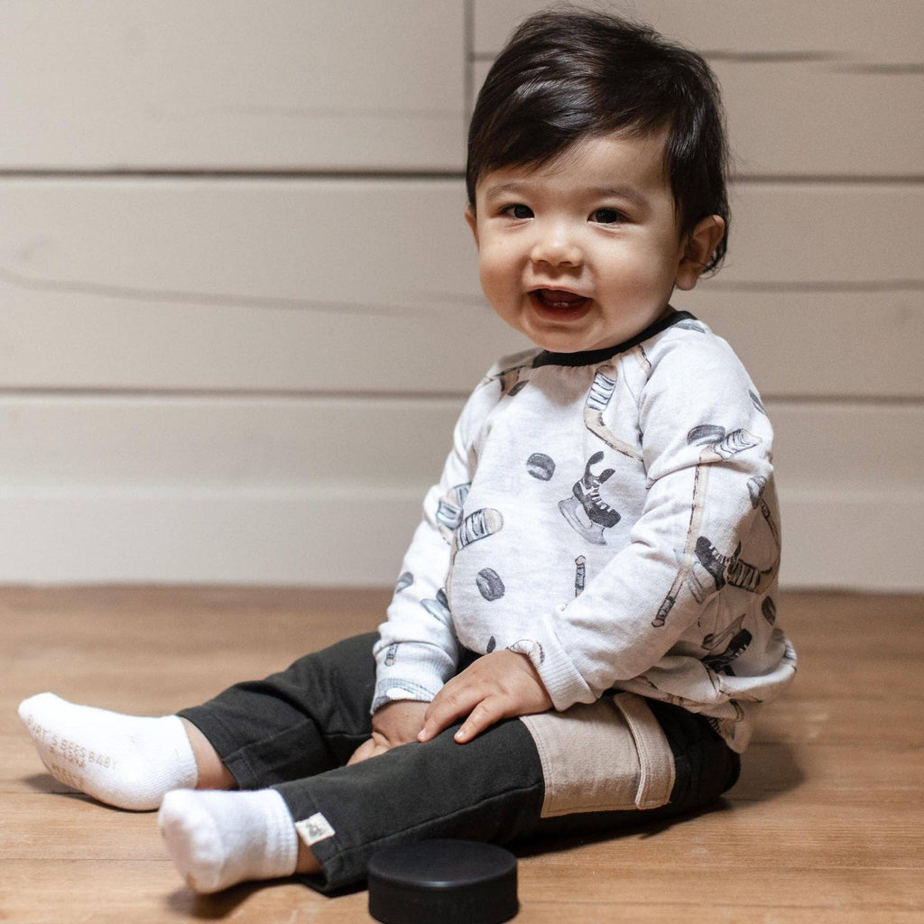 Smiling baby sitting on a wooden floor wearing Burt's Bees Baby - Hockey Season Sweatshirt & Pants Set with hockey-themed prints and soft neutral tones, styled for cozy everyday wear.