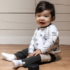 Smiling baby sitting on a wooden floor wearing Burt's Bees Baby - Hockey Season Sweatshirt & Pants Set with hockey-themed prints and soft neutral tones, styled for cozy everyday wear.