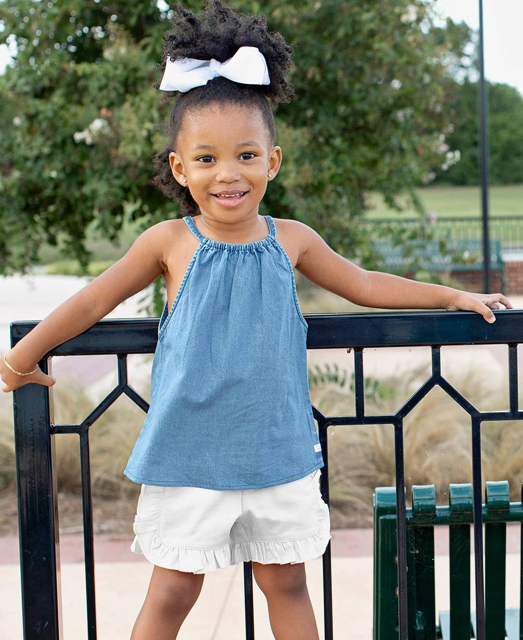 Toddler girl wearing RuffleButts + RuggedButts - Toddler Girls White Ruffle Trim Woven Shorts: White, styled with a sleeveless blue top, standing outdoors with a big white bow in her hair.
