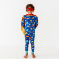 A happy child wearing vibrant blue pajamas with colorful superhero and space motifs, a red superhero mask, and a matching red and yellow cape showcasing Kiki + Lulu - Cozy & Courageous Superhero Kids Pajamas.