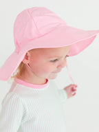 Young child wearing a pink RuffleButts + RuggedButts - Kids Sun Protective Hat with wide brim, soft pastel clothing in a bright, minimalist setting, stylish kids clothing.