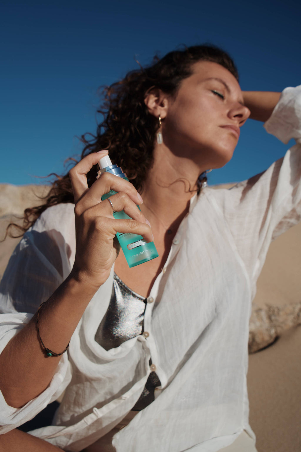 Woman outdoors in bright sunlight applying True Ocean - True Ocean: Body Spray - 3.4oz. with eyes closed, wearing light white blouse and silver top, evoking fresh coastal vibes.