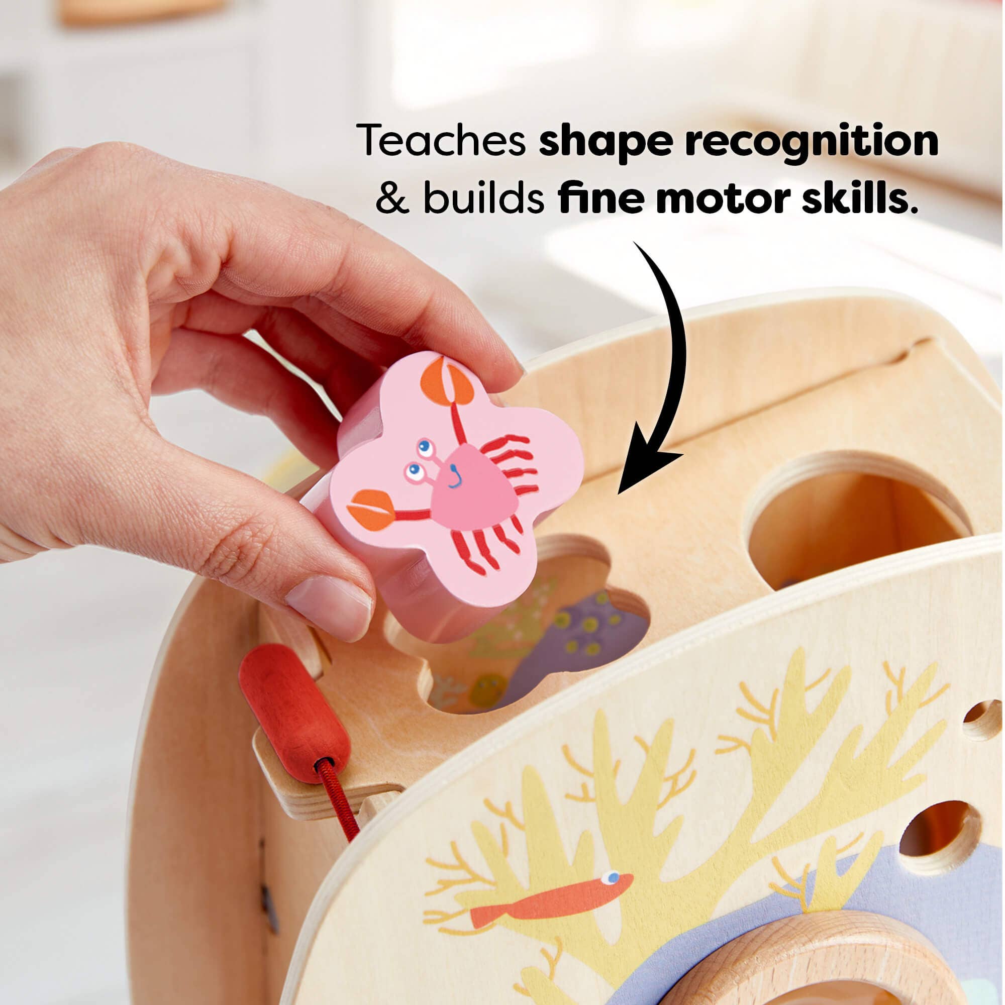 Hand placing a pink crab-shaped wooden piece into the matching hole on an Underwater World Wooden Shape Sorter, designed to teach shape recognition and fine motor skills.