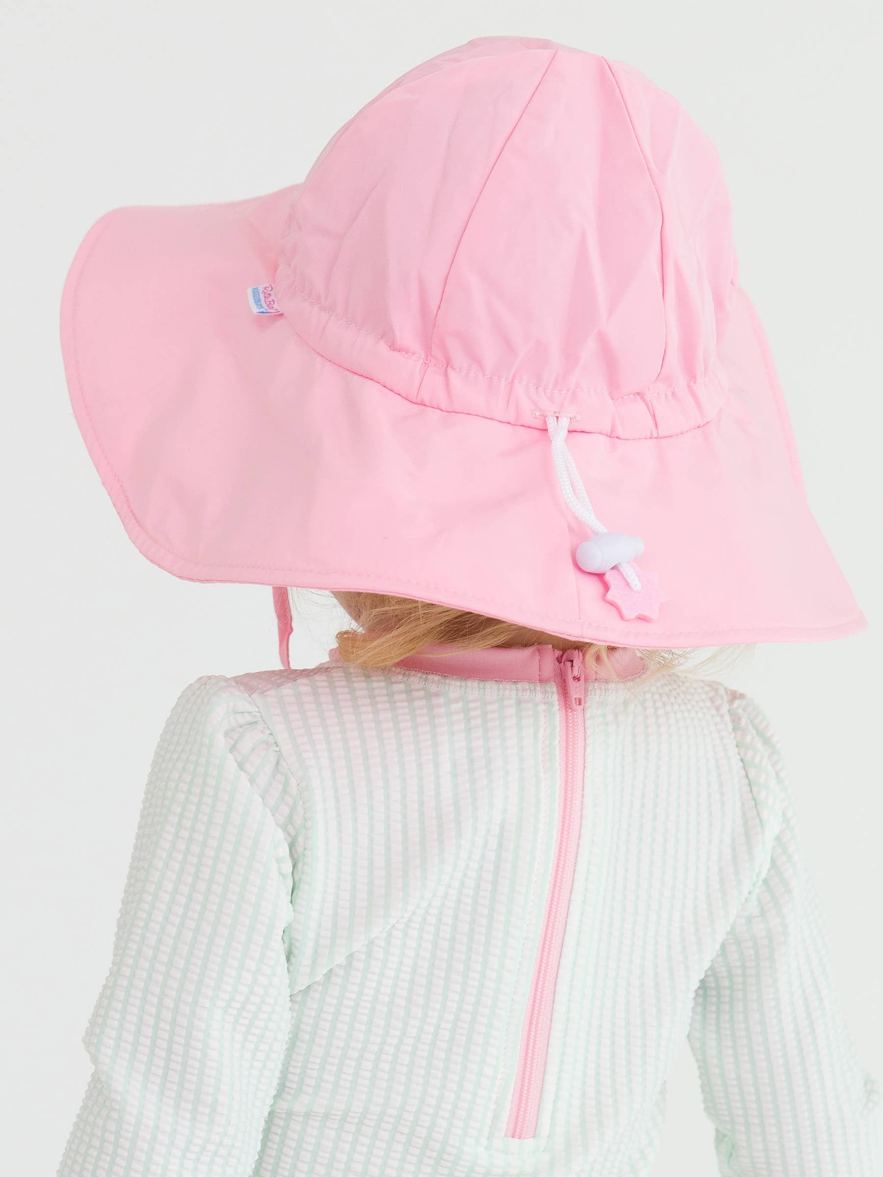 Back view of a child wearing a light pink RuffleButts + RuggedButts - Kids Sun Protective Hat with adjustable chin strap, paired with a textured light-colored outfit, showcasing stylish kids clothing from a curated children’s boutique.