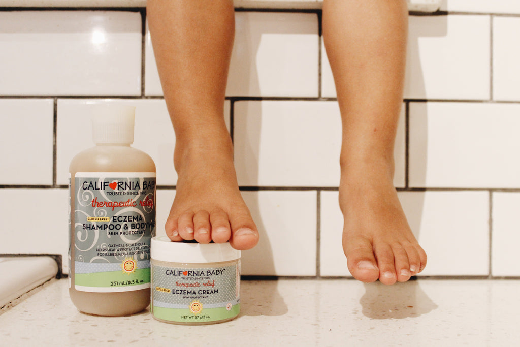 Child’s feet above white tiled bath floor with California Baby - Eczema Cream + Wash Bundle products placed nearby, highlighting cozy baby essentials for sensitive skin care.