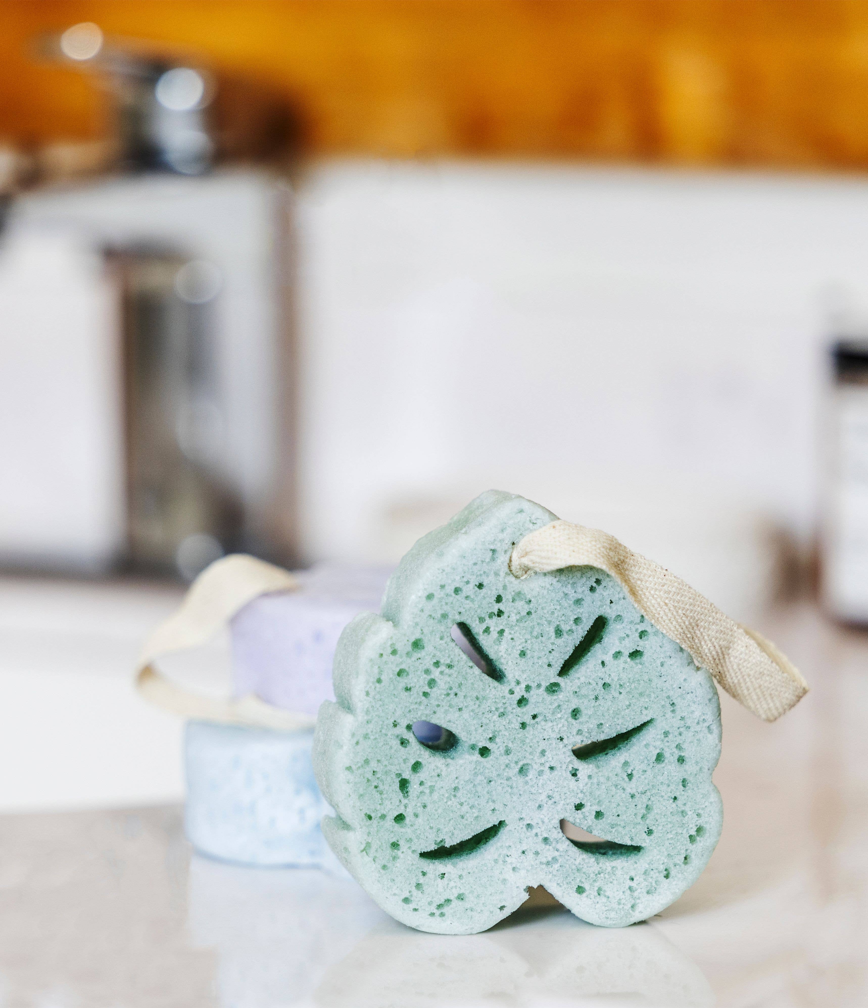 Lemon Lavender - In The Buff Super Sudsy Soap Sponge in light green with natural texture and attached loop, styled in a cozy modern bathroom setting.