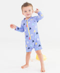 Toddler boy wearing the RuffleButts + RuggedButts - Baby Boys Down By The Bay Long Sleeve One Piece Rash Guard Swimsuit: Blue with colorful sailboat prints, standing barefoot on a white background.