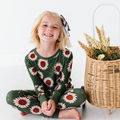Smiling young girl sitting cross-legged wearing Kiki + Lulu - Rustic Blooms Ribbed Toddler/Big Kid Pajamas with large floral sunflower prints on a green background beside a wicker basket with dried flowers.