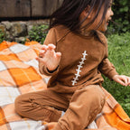 Child wearing a brown sweatshirt outfit with a football design, sitting on an orange plaid blanket outdoors.