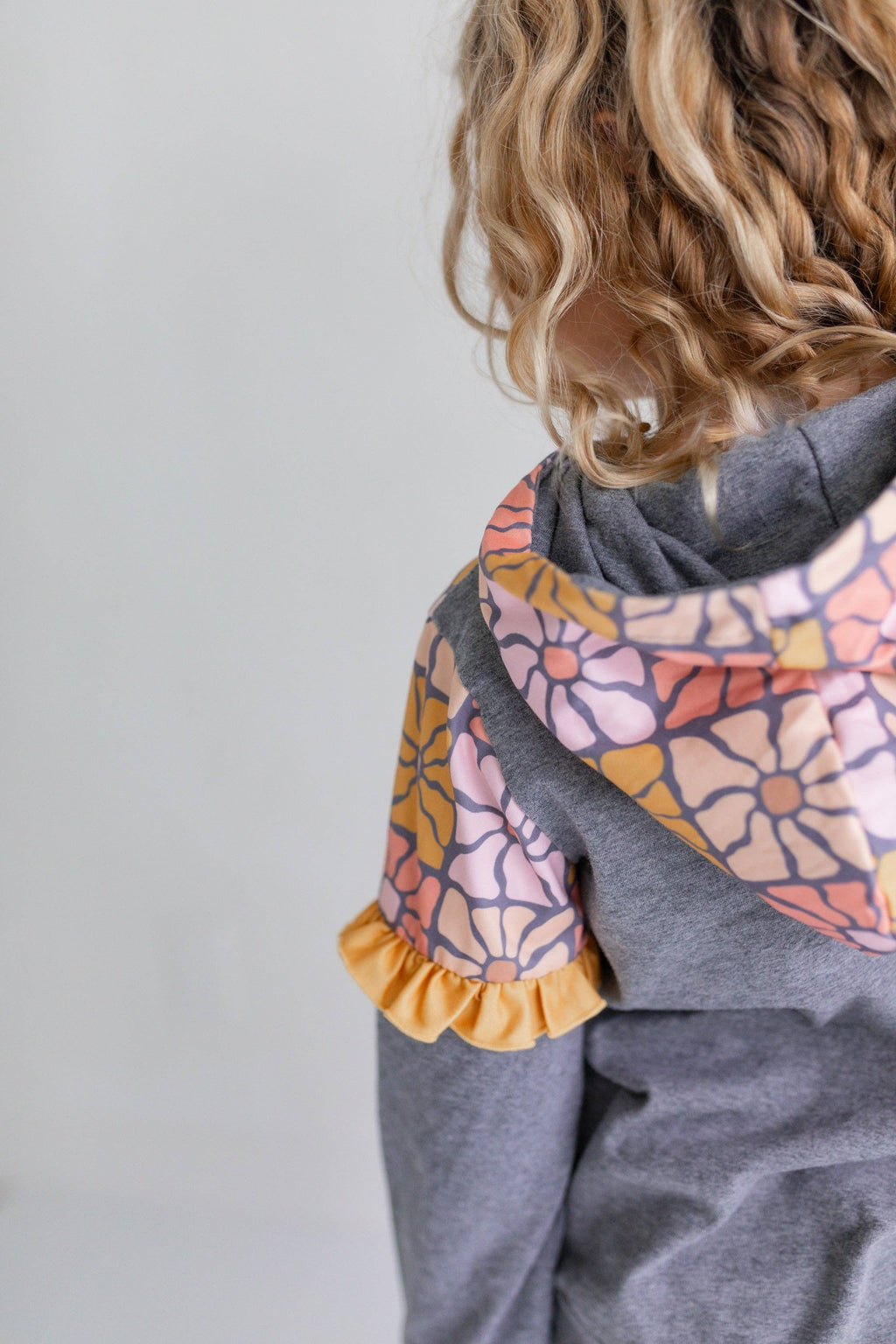 A child wearing a gray hoodie with a colorful floral-patterned hood and matching patterned sleeve cuff, shown from the back against a light gray background.