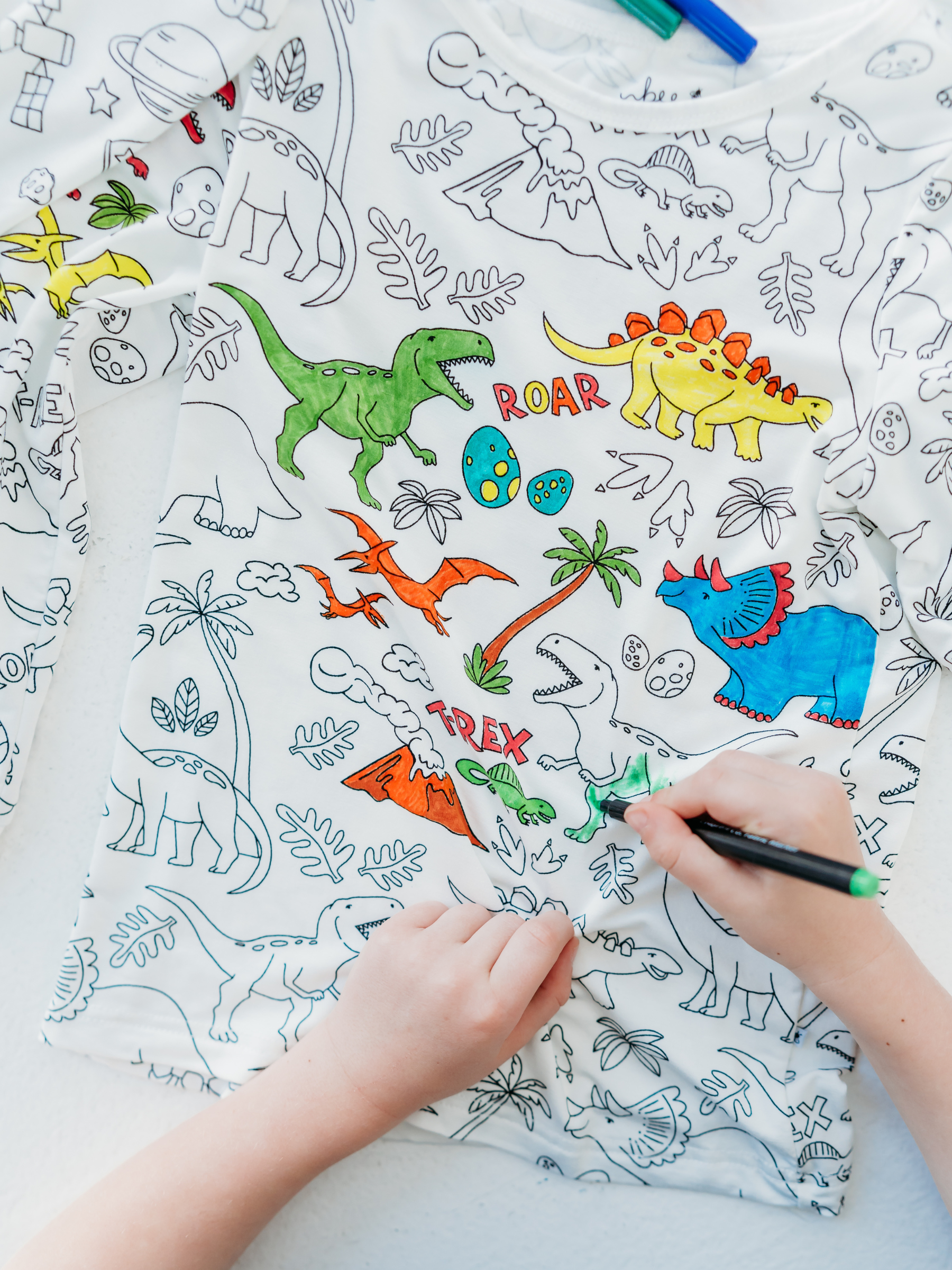 Child coloring dinosaur-themed pajama with green fabric marker, demonstrating use of Bee Colorful Co. - Fabric Markers Set for Customizing Coloring Pajamas from a curated children’s boutique focused on stylish kids clothing and mom life essentials.
