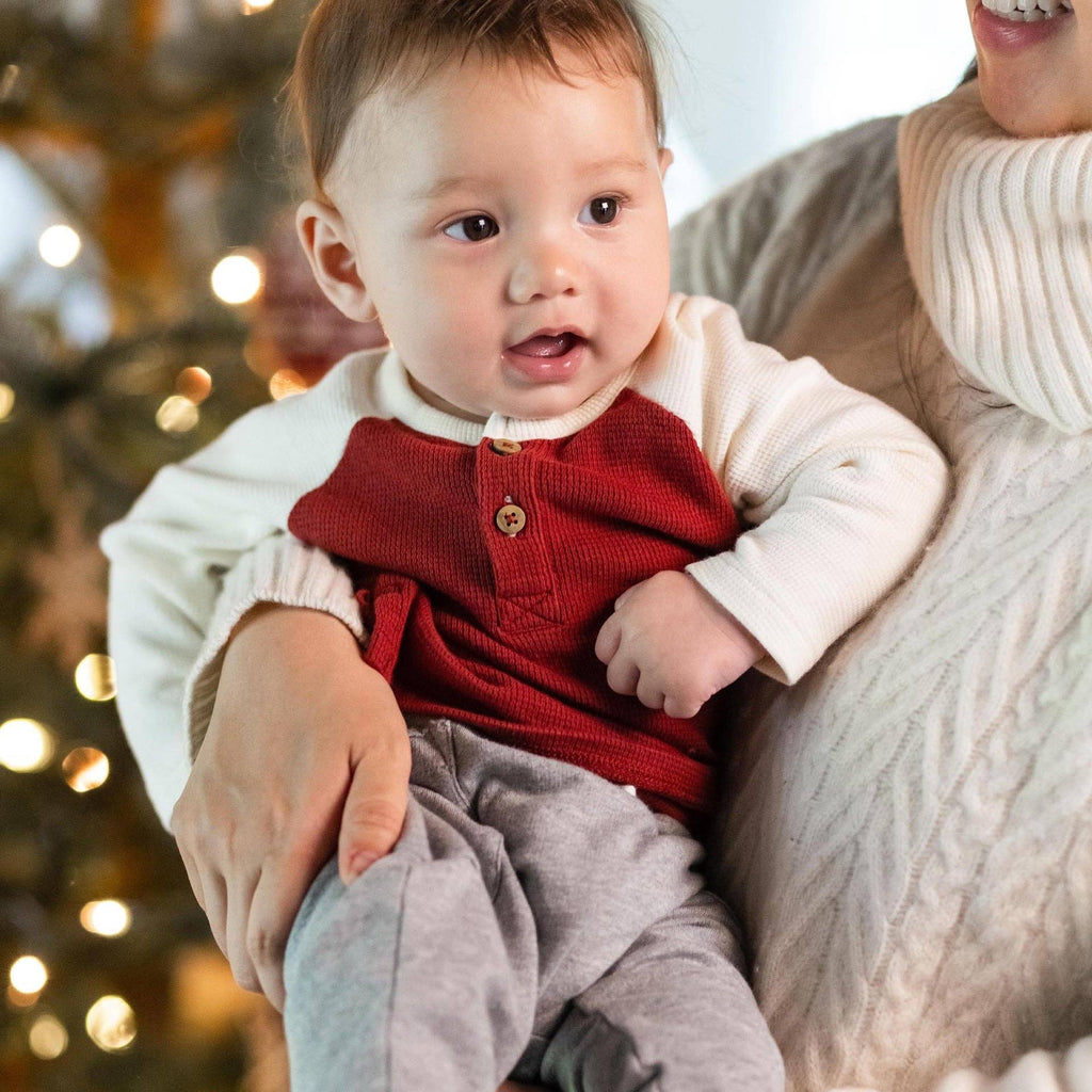Baby boy wearing Burt's Bees Baby - Thermal Organic Boy Tee & Pants Set - Brick Red, held by parent near decorated holiday tree in cozy home setting.